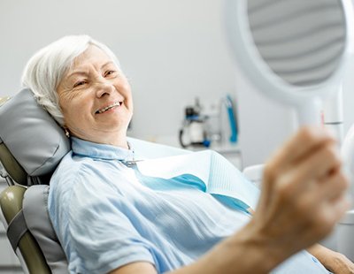 older lady sitting in dental chair and smiling into hand mirror after dental bonding treatment