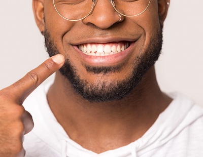 An up-close image of a young man wearing glasses and a white hoodie, pointing to his newly straightened smile