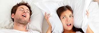 Woman covering ears in bed with snoring man who needs sleep apnea therapy