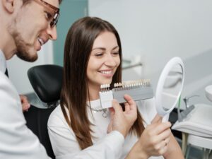 Dentist holding veneers up to patient while she smiles into handheld mirror.
